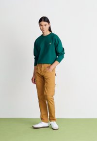 Grüner Sweatshirt mit gerippten Bündchen und kleinem Logo, kombiniert mit beige Cargo-Hosen mit Seitentaschen, getragen mit weißen Turnschuhen.