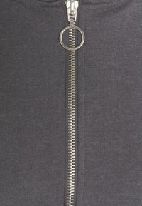 Giacca grigia con zip metallici e un cursore circolare. Il tessuto è texturizzato, aderente e ribbed lungo i bordi.