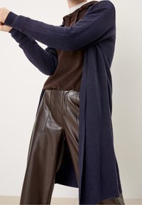 Langer, marineblauer, gerippter Cardigan aus strukturiertem Strick, über einem braunen Hoodie und braunen Kunstlederhosen getragen, mit dehnbaren Ärmeln.