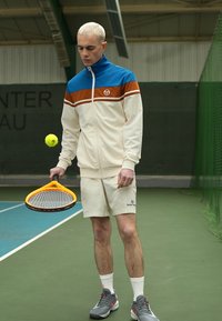 Un chándal ligero en crema con detalles en azul y naranja, combinado con pantalones cortos beige. El modelo sostiene una raqueta de tenis naranja y una pelota amarilla.