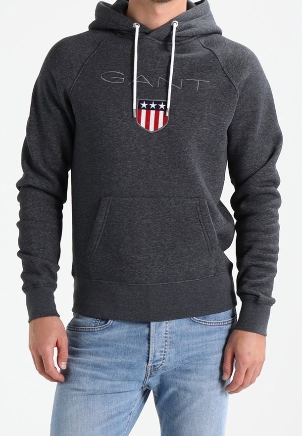 Homme portant un sweat à capuche gris foncé avec cordons blancs, une poche avant et un logo GANT avec un emblème de bouclier rouge, blanc et bleu.