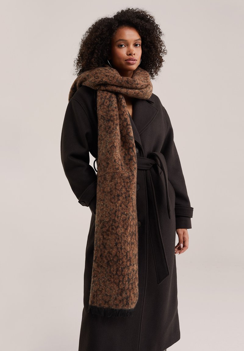 Femme aux cheveux bouclés portant un long manteau noir ceinturé et une épaisse écharpe brune à motifs enroulée autour du cou.