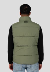 Mann mit einer hellgrünen gepolsterten Weste mit dem Text "WRONG FRIENDS" auf dem Rücken, über einem schwarzen Langarmshirt und hellblauen Jeans, von hinten zu sehen.