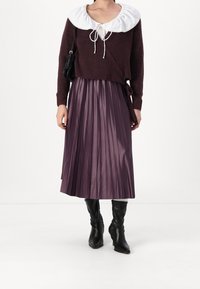 Kvinna i en mörklila omslagströja med en stor vit krage, en glänsande plisserad lila midikjol, svarta klackkängor och en svart axelväska.