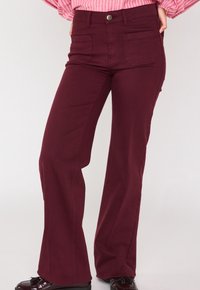 Pantaloni a fiamma bordeaux in un morbido tessuto elasticizzato. Presentano tasche frontali e posteriori, chiusura con bottone e dettagli cuciti lungo le cuciture.