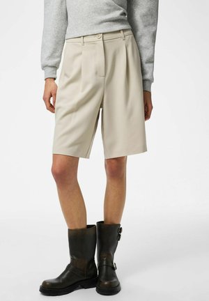 Shorts beige ajustés avec plis avant, taille haute et poches latérales, associés à des bottines en cuir noir avec boucles.