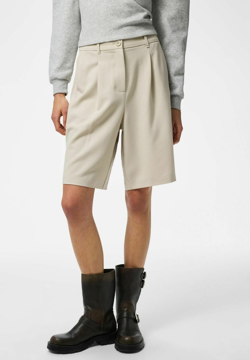 Shorts beige ajustés avec plis avant, taille haute et poches latérales, associés à des bottines en cuir noir avec boucles.
