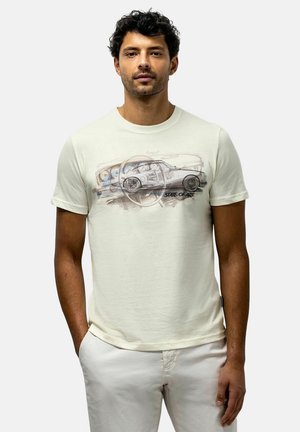 Man met krullend donker haar die een gebroken witte t-shirt draagt met een schets van een vintage auto en stuurwiel en tekst "state of art", staand met handen in de zakken.