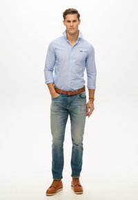 Camicia button-up a righe azzurre, maniche arrotolate, con logo blu scuro. Indossata con jeans blu sbiaditi e scarpe in pelle marrone, cintura casual.
