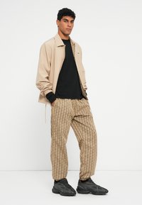 Giovane uomo che indossa pantaloni beige con motivo, giacca beige sopra una maglietta nera e sneakers nere, in piedi davanti a uno sfondo bianco semplice.