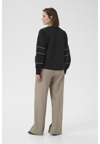 Blouse noire à manches bouffantes, ornée d'une bordure à motif blanc. Associée à un pantalon large beige clair et des chaussures noires.