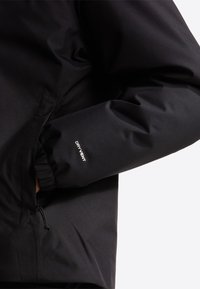 Άτομο που φοράει μαύρο μπουφάν με ελαστικό μανίκι και κείμενο "DRYVENT" στο μανίκι, με το χέρι του μέσα στην πλαϊνή τσέπη.