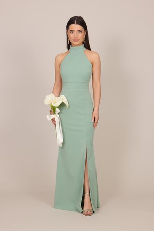 Frau in einem ärmellosen salbeigrünen Kleid, die weiße Calla-Lilien hält, die mit einem weißen Band gebunden sind, stehend vor beigem Hintergrund.