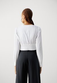 White smocked back blouse with long sleeves, featuring a round neckline and a textured finish, paired with high-waisted black trousers.
