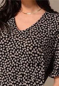 Černá blůza s malým bílým květinovým vzorem. Má výstřih do V a krátké rukávy. Hladký materiál s jemným skládáním.