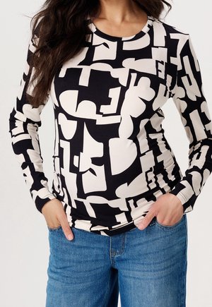 Femme portant un haut ajusté à manches longues noir et blanc avec un motif abstrait, un jean bleu, les mains dans les poches, sur un fond uni.