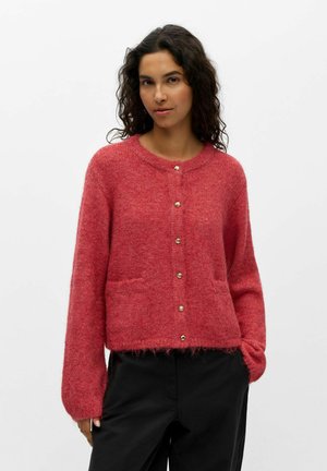 Femme aux cheveux bouclés et foncés, portant un cardigan rouge boutonné en matière duveteuse et un pantalon noir, debout devant un fond blanc uni.