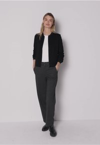 Musta pommikenttä takki valkoisen topin päällä, yhdistetty harmaisiin housuihin. Malli seisoo käsivartensa taskuissa, mustat kengät jalassa. Yksinkertainen muotoilu.