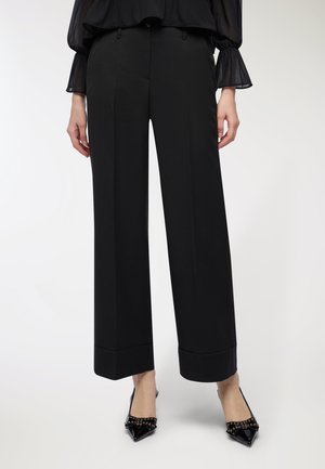 Donna che indossa pantaloni neri a gamba larga e décolleté nere a punta con fiocchi borchiati, in piedi su uno sfondo bianco semplice.