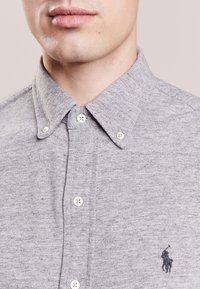 Szara koszula z guzami, uszyta z teksturowanej tkaniny, z klasycznym kołnierzykiem, białymi guzikami oraz małym haftowanym logo na piersi.