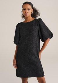 Rochie neagră cu textură, cu o siluetă pătrată, decolteu rotund și mâneci bufante. Țesătura are un model subtil.