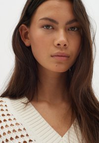 Gestrickter Pullover in Off-White mit tiefem V-Ausschnitt, der eine Mischung aus offenen und gerippten Texturen aufweist, getragen von einem Model mit langen, dunklen Haaren.