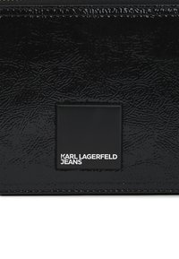 Materiale in pelle nera testurizzata con un'etichetta quadrata nera liscia impressa con la scritta "KARL LAGERFELD JEANS" in testo bianco.