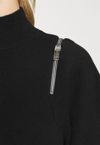 Pull noir en maille côtelée avec col montant et détail de fermeture éclair argentée sur l'épaule, porté par une personne aux cheveux blonds tirés en arrière.