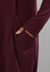 Burgundowy detal odzieżowy z miękką teksturą, długimi rękawami i boczną kieszenią o luźnym fasonie.