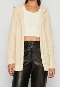 Žena oblečená v krémovém pleteném kardiganu s kapucí, bílém ribbovaném zkráceném topu a černých kožených kalhotách s vysokým pasem na jednobarevném pozadí.