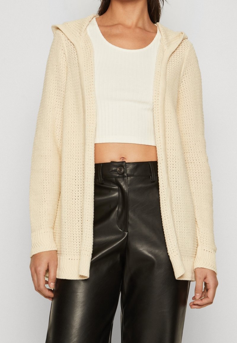 Žena oblečená v krémovém pleteném kardiganu s kapucí, bílém ribbovaném zkráceném topu a černých kožených kalhotách s vysokým pasem na jednobarevném pozadí.