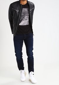 Man som bär svart läderjacka, grafisk svart T-shirt, mörkblå jeans och vita sneakers, står mot vit bakgrund.