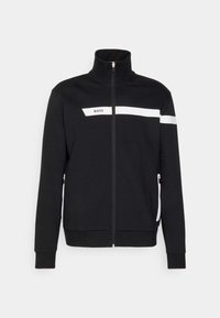 Veste noire zippée avec col montant, bande blanche horizontale sur la poitrine et le bras, logo "BOSS" sur la poitrine gauche, et poches latérales zippées.
