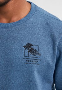 Blauwe sweatshirt van textiel met structuur. Heeft een ronde hals, geribde manchetten en een geborduurd ontwerp met bomen en bergen.