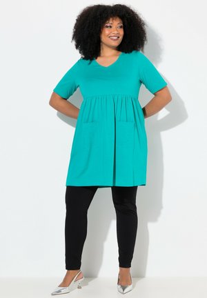 Een turquoise tuniek met korte mouwen, voorzien van een V-hals, geplooide taille en twee frontzakken, gecombineerd met zwarte leggings en zilveren hakken.