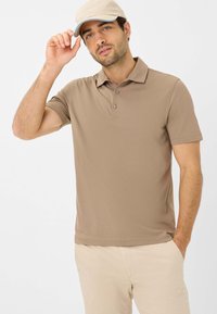Polo shirt marrone con colletto, tre bottoni e maniche corte, abbinato a pantaloni beige. Modello che indossa un cappellino beige chiaro.