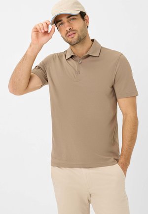 Bruine polo met een kraag, drie knopen en korte mouwen, gecombineerd met beige broeken. Model draagt een lichtbeige pet.