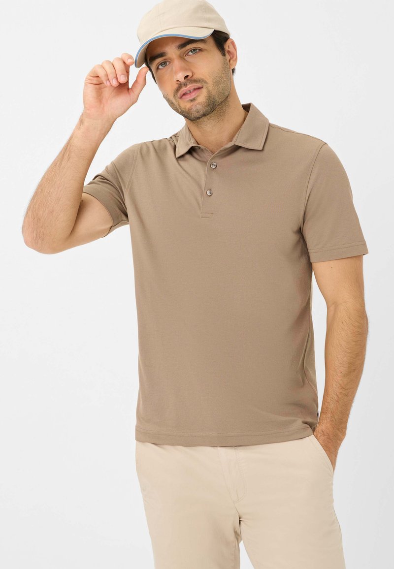 Polo shirt marrone con colletto, tre bottoni e maniche corte, abbinato a pantaloni beige. Modello che indossa un cappellino beige chiaro.