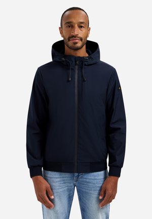 Man draagt een marineblauwe capuchonjas met rits aan de voorkant en trekkoorden, gecombineerd met lichtblauwe spijkerbroek, kijkend naar voren tegen een witte achtergrond.