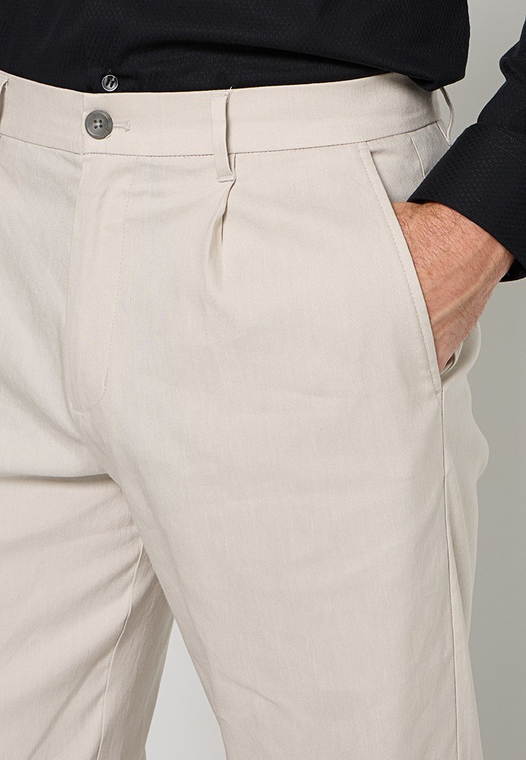 Mann mit beigen Hosen, die Hand in der rechten Tasche, und einem schwarzen Langarmhemd, das eingesteckt ist und Knopf sowie Gürtelschlaufen zeigt.