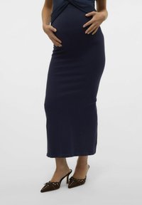 Robe de maternité en ribbed marine avec un haut noué, présentant une silhouette ajustée et une longue jupe fluide. Accentuée par des escarpins bruns à bout pointu.