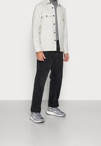 Chemise boutonnée gris clair avec deux poches poitrine, associée à un jean gris foncé et des baskets grises. La chemise a une coupe décontractée et un col pointu.