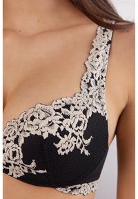 Soutien-gorge noir avec des détails en dentelle beige complexe sur les bretelles et les bonnets, avec un tissu texturé et un motif floral le long des bords.