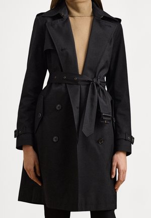Femme portant un trench-coat noir à double boutonnage avec une ceinture nouée, des poignets boutonnés et des larges revers, par-dessus un pull col roulé beige.