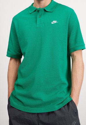 Muž v zelené polo košili Nike a tmavých šortkách Nike s rukama v kapsách na jednoduchém pozadí.