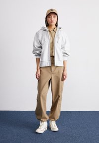 Lys grå hettejakke med store ermer, kombinert med khaki cargo-bukser. Hvite høyhælte sneakers fullfører antrekket. Beige caps.