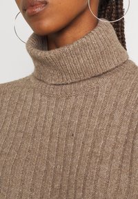 Béžový žebrovaný rolákový svetr vyrobený z texturovaného pleteného materiálu. Má vysoký límec a viditelné šití.