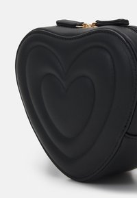 Czarna skórzana torebka w kształcie serca z wytłaczanym wzorem serca i złotym zjazdem, na tle jednolitego białego tła.