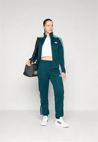 Miętowy dres z białymi paskami po bokach, kurtka na suwak i spodnie z elastyczną talią. Całość dopełniają białe buty i czarna torba sportowa.