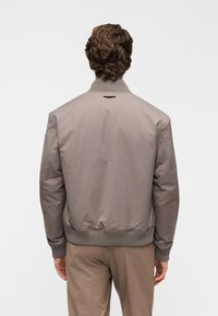 Ανοιχτό καφέ bomber μπουφάν με ριμπ γιακά και μανσέτες, με λεία υφή και μικρή λεπτομέρεια λογοτύπου στην πίσω πλευρά.